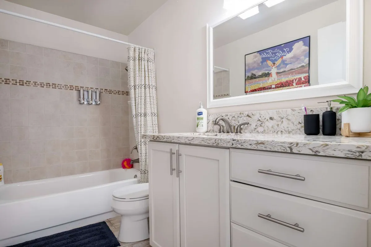A beautifully appointed bathroom featuring a white bathtub, granite countertop, framed mirror, stylish decorative artwork, and a green plant on the counter. The space is filled with natural light and has a calming, fresh ambiance that invites relaxation.