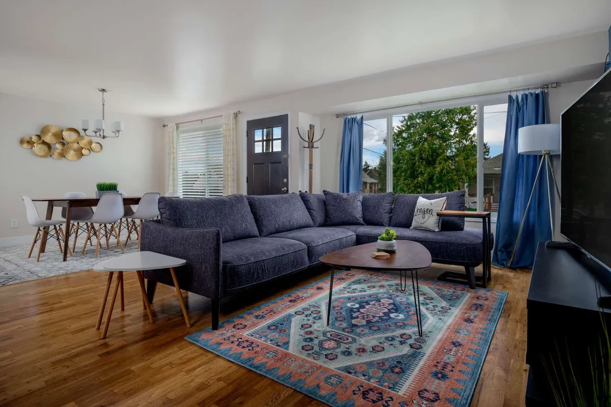 The image showcases a sunlit living and dining area with a plush blue sectional sofa, a stylish wooden coffee table, a large window that fills the room with natural light, and a spacious dining table—creating an open, cozy, and social atmosphere.