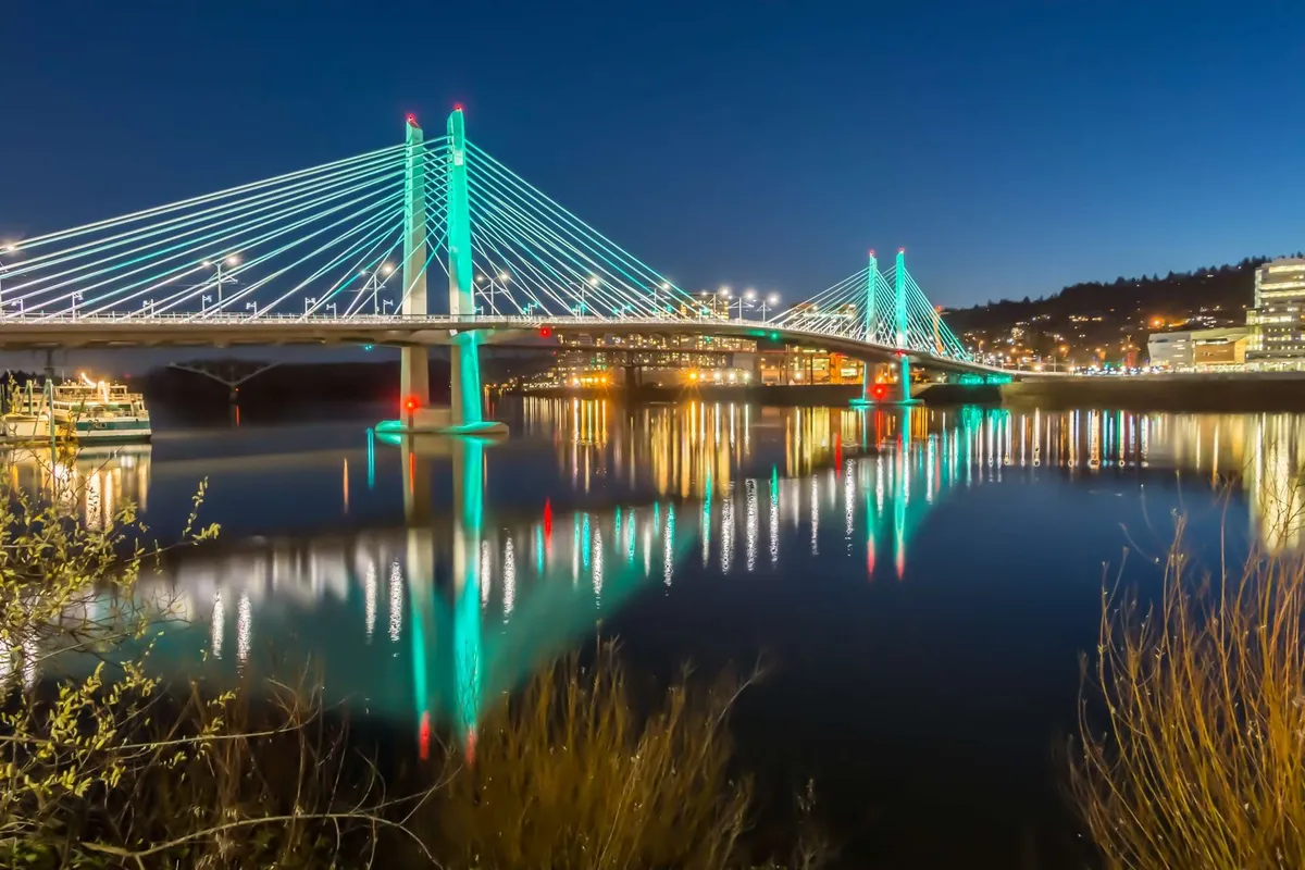 A panoramic evening view featuring a lit suspension bridge, sparkling city lights reflected in the water, and the lively ambiance of a riverside setting. The scene captures the vibrancy and accessibility of nearby city amenities, inviting viewers to explore local attractions beyond the property.