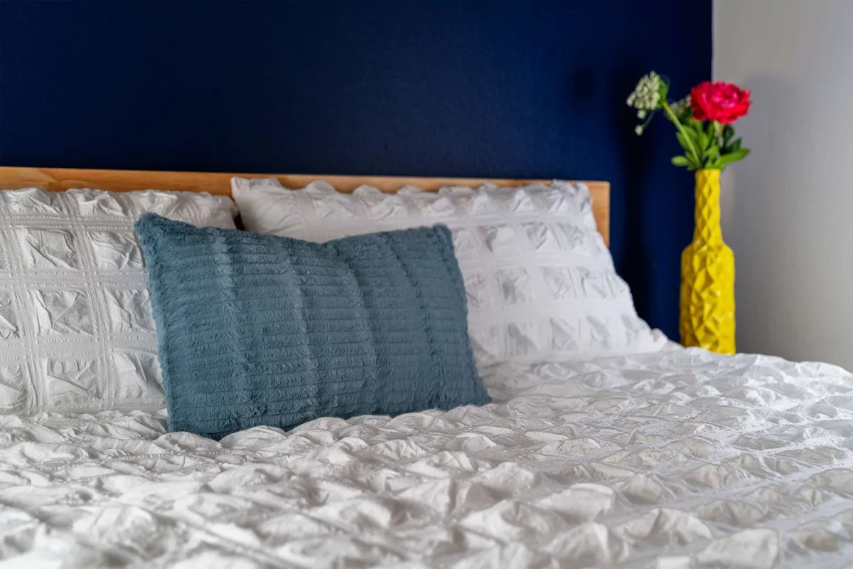 A tranquil bedroom scene featuring a bed with crisp white bedding, accented by a teal pillow and a yellow vase with fresh flowers. The soft natural light and uncluttered decor evoke a peaceful, restful atmosphere.