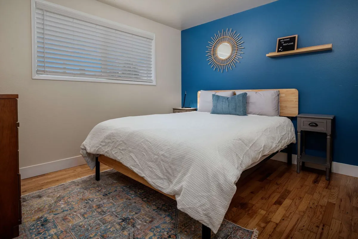 This image showcases a serene bedroom featuring a plush bed with crisp white bedding, a calming blue accent wall, a decorative mirror above the headboard, and a nightstand with a glowing lamp. The overall vibe is one of tranquility and restful comfort, ideal for unwinding after a busy day.