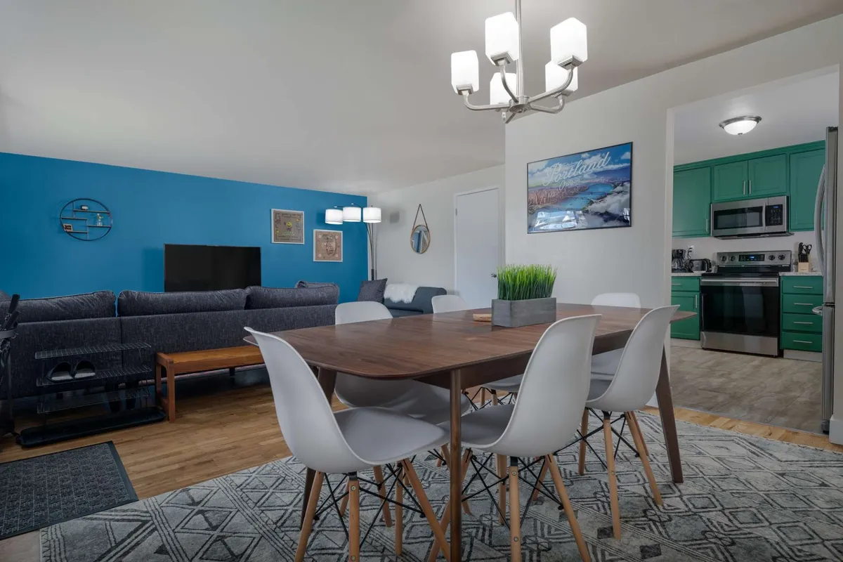 A bright, open-concept living and dining space featuring a dining table with white chairs, a cozy sofa area, a blue accent wall, and the entrance to the kitchen. Decorative artwork adds a modern touch, and the layout encourages easy flow between spaces.