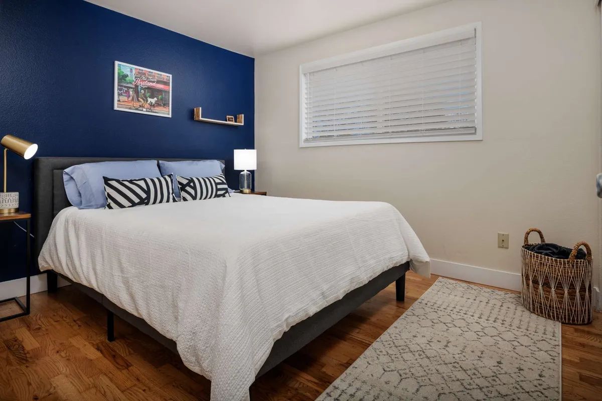 A peaceful bedroom highlighted by a large bed with crisp white bedding, a bold blue accent wall, decorative artwork, and a nightstand with a softly lit lamp. The ambiance is both calm and welcoming, suggesting a space designed for relaxation and deep sleep.